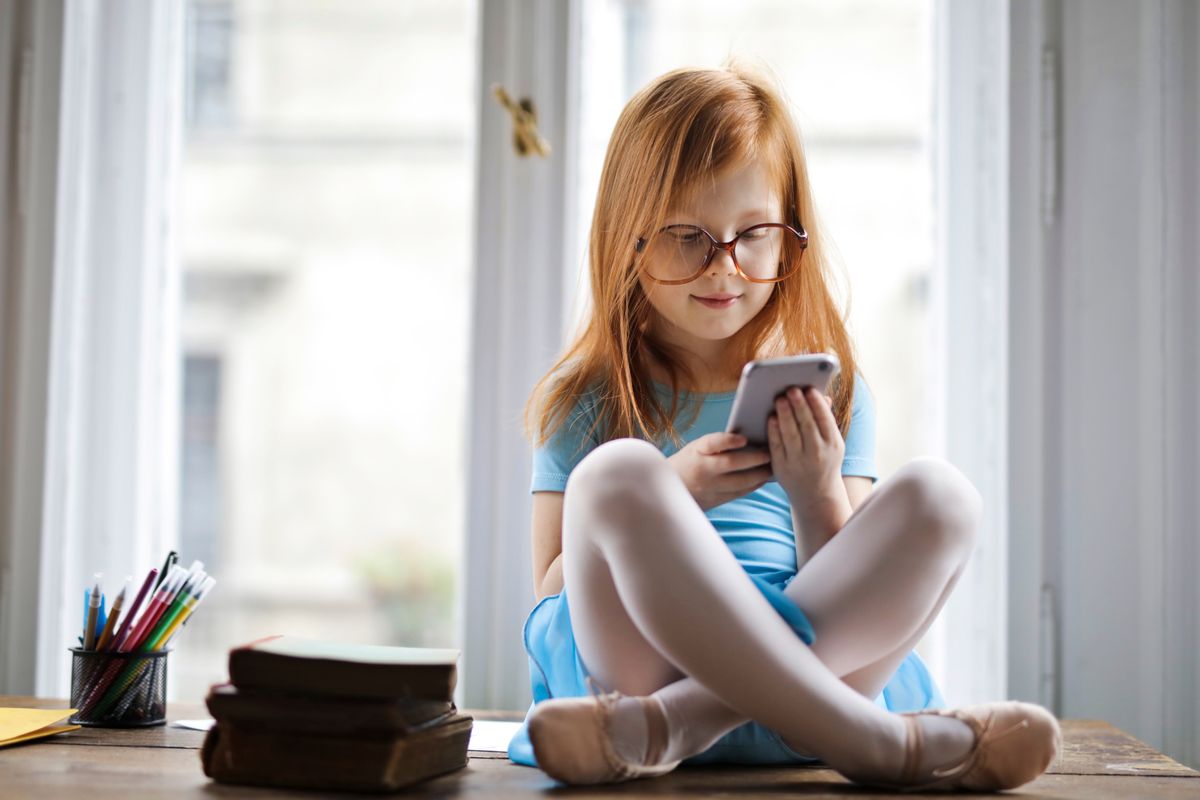 Adorabile ragazza dai capelli rossi con gli occhiali che usa uno smartphone all'interno di una stanza accogliente, seduta accanto ai libri. - Foto di Andrea Piacquadio su Pexels