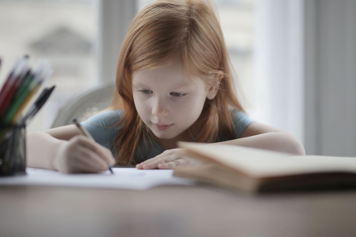 Adorable positive preschool child with red hair in casual wear sitting at table with book and pens or pencils while making exercise on paper sheet against blurred window - Photo by Andrea Piacquadio on Pexels
