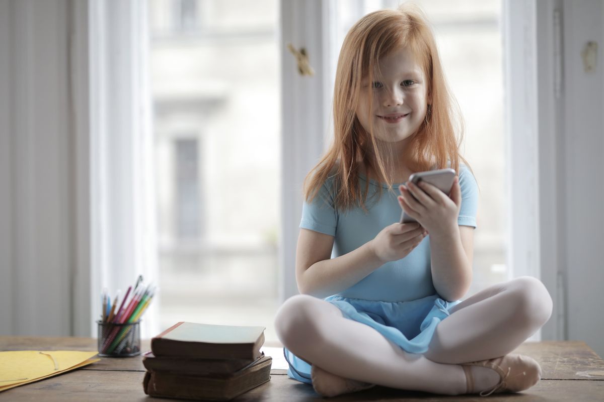A cheerful young girl in a blue dress sitting indoors by a window holding a smartphone. - Photo by Andrea Piacquadio on Pexels