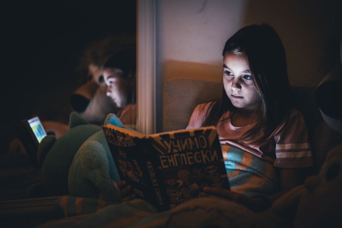 A young girl reads a book under dim light, reflecting a quiet bedtime atmosphere. - Photo by Boris Hamer on Pexels