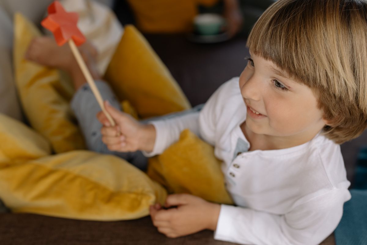 Cheerful child smiling while holding a red star-shaped wand surrounded by yellow pillows indoors. - Photo by cottonbro studio on Pexels