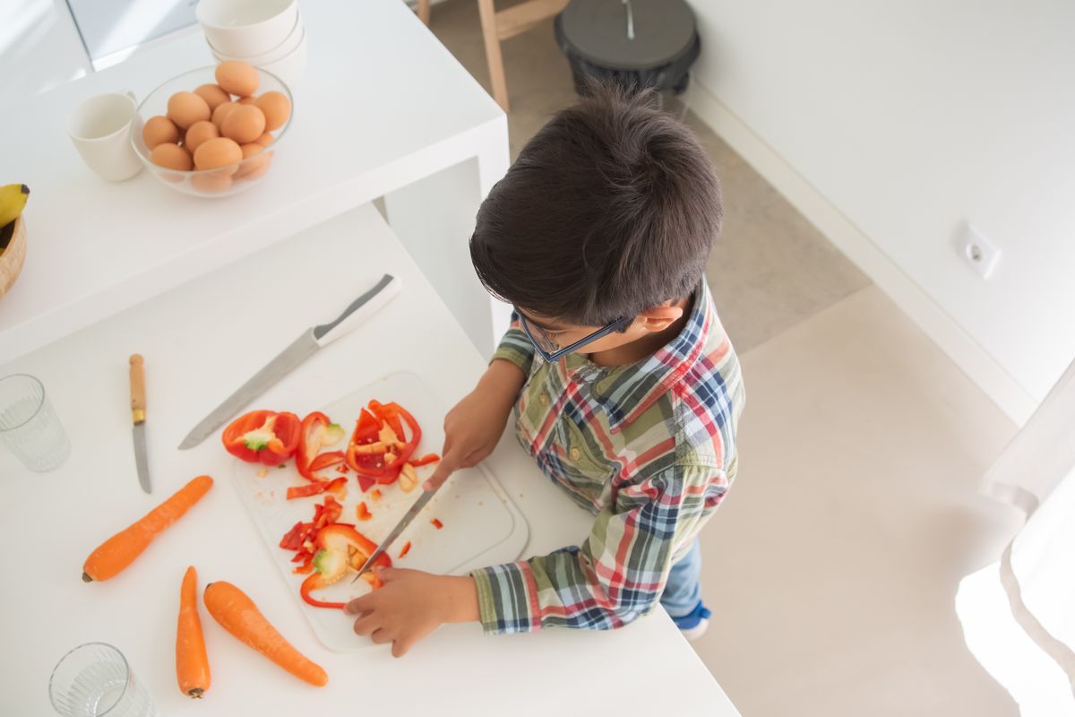A child in a checkered shirt slices vegetables on a chopping board in a modern kitchen. - Photo by Kampus Production on Pexels