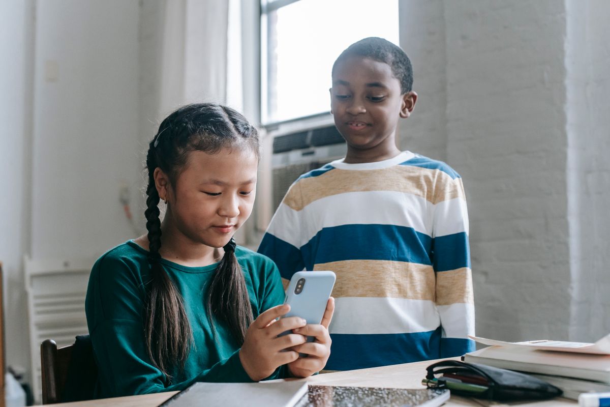 Happy little African American boy standing near Asian classmate girl using smartphone sitting at desk during break - Photo by Katerina Holmes on Pexels