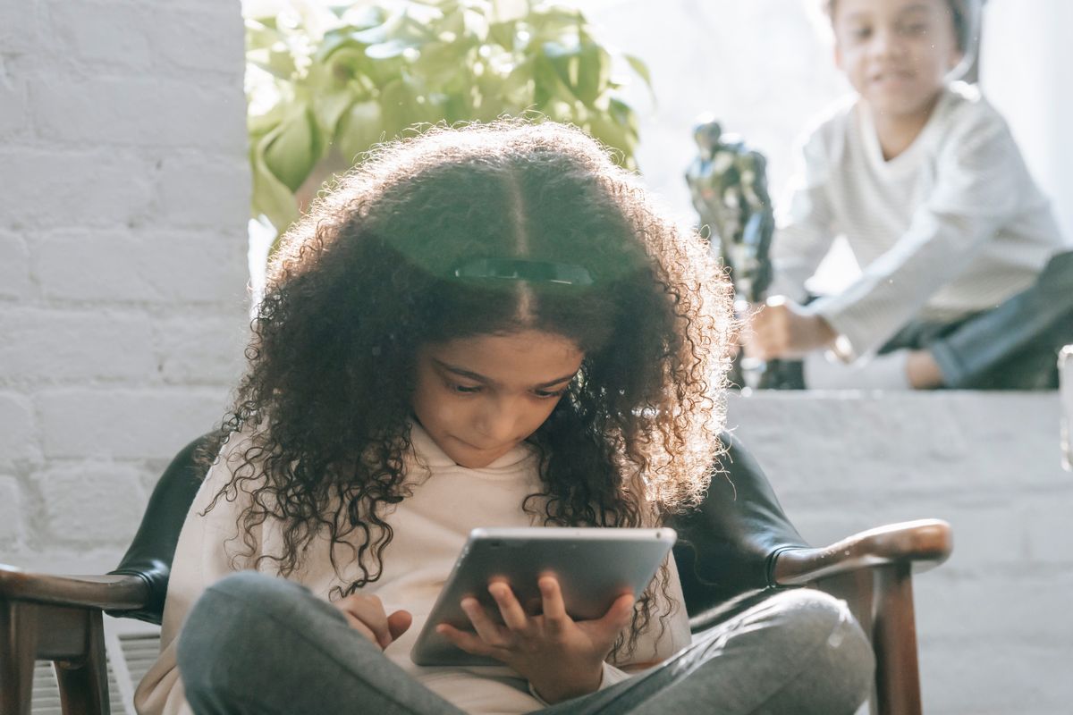 Concentrated African American kid with long curly hair watching video on tablet sitting on comfortable armchair near crop smiling little brother playing with toy - Photo by Marta Wave on Pexels