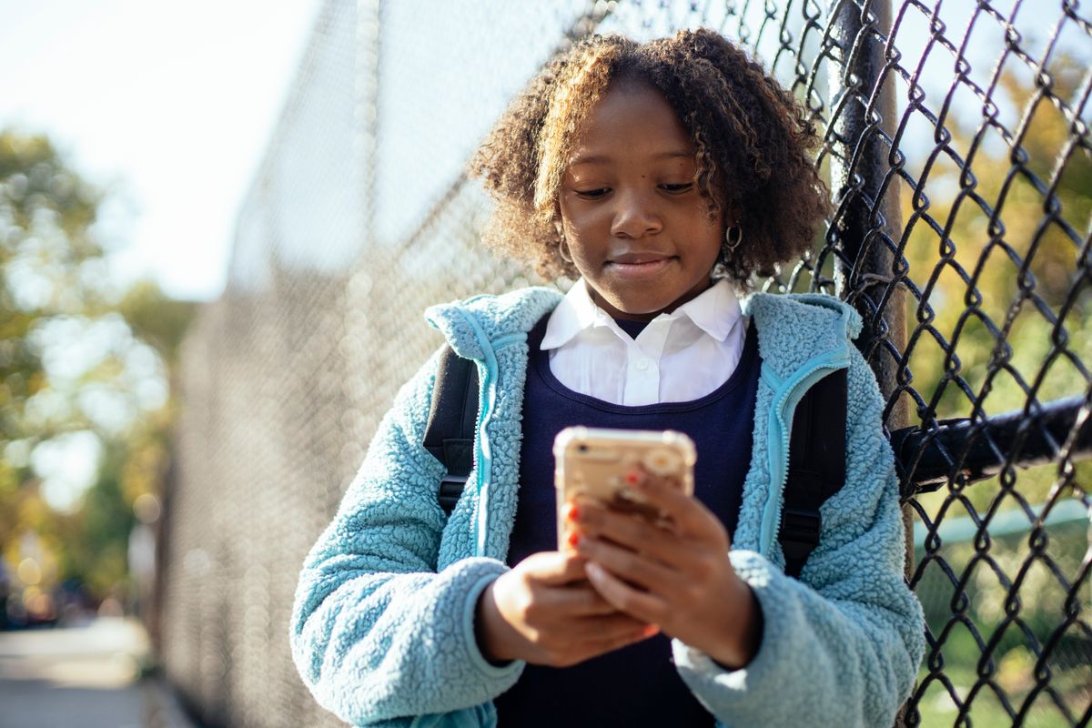 Positive African American schoolgirl in blue jacket browsing mobile phone while standing near wire net fence on sunny autumn day Positives afroamerikanisches Schulmädchen in blauer Jacke, das an einem sonnigen Herbsttag in der Nähe eines Maschendrahtzauns steht und auf dem Handy surft