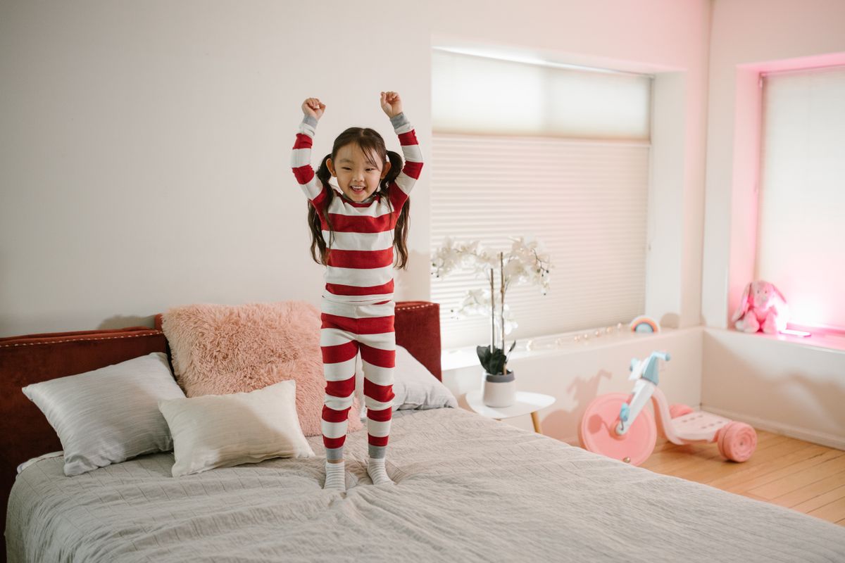 A joyful child in striped pajamas jumping on a bed in a bright and cozy bedroom setting. - Photo by RDNE Stock project on Pexels