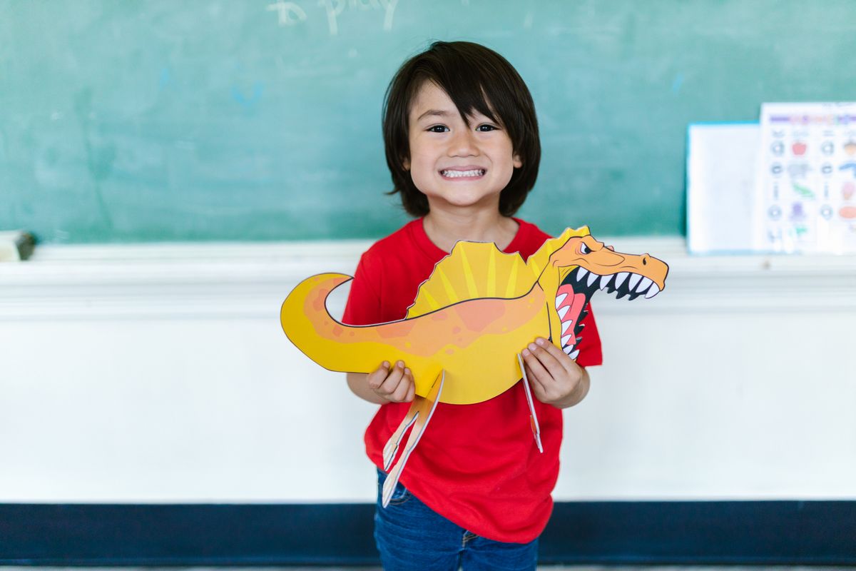 A cheerful child holds a colorful dinosaur cutout in a classroom setting. - Photo by RDNE Stock project on Pexels